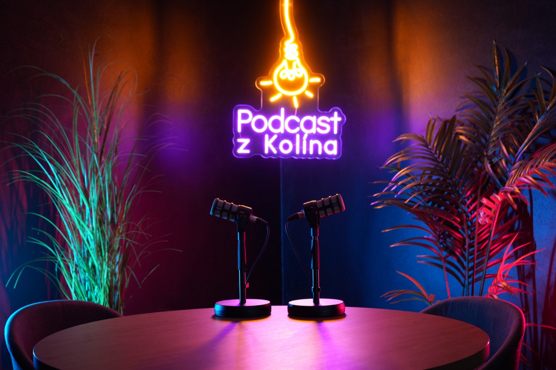 Podcastové studio A s neonovým logem Podcast z Kolína a dvěma mikrofony na kulatém stole